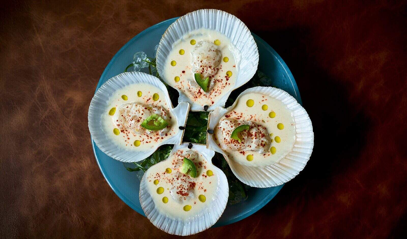 Four scallop shells filled with creamy sauce, garnished with green herbs, red seasoning, and drops of oil, arranged on a blue plate—discover this hidden gem at 1 Hotel in South Lake Union.
