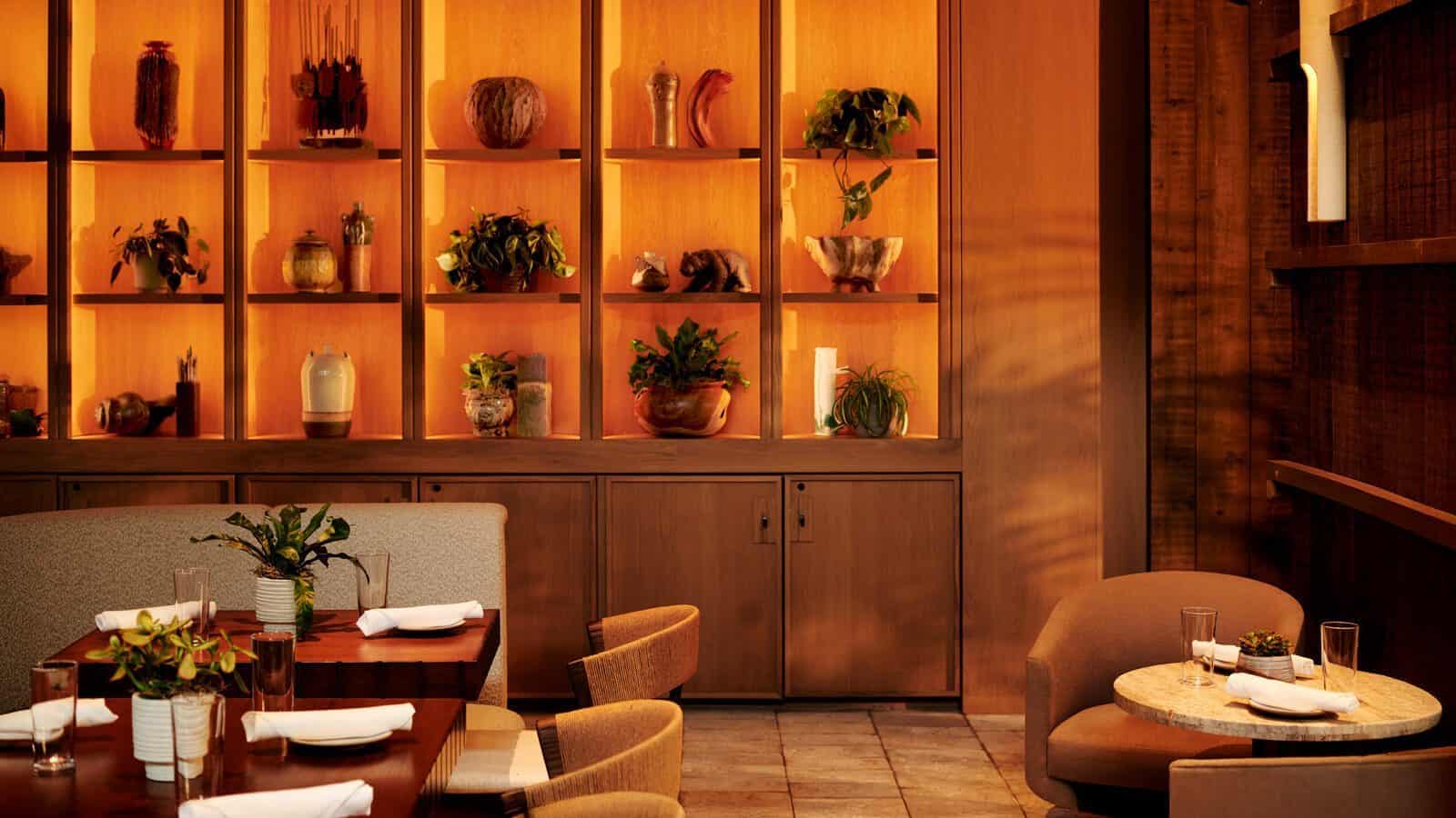 A warmly lit restaurant interior at 1 Hotel in South Lake Union, a hidden gem with wooden shelves displaying pottery and plants, and tables set with glasses, plates, and napkins.