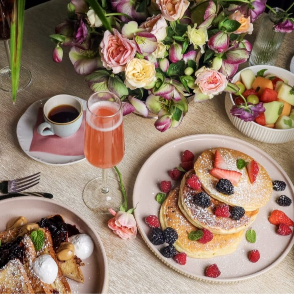 A table set with pancakes topped with berries, a fruit bowl, French toast, a cup of coffee, a pink drink, and a bouquet of flowers.