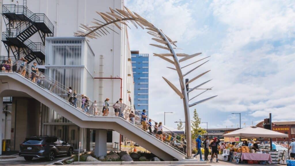 People walk up an outdoor staircase beside a large fishbone-shaped metal sculpture, with market stalls and buildings visible in the background.