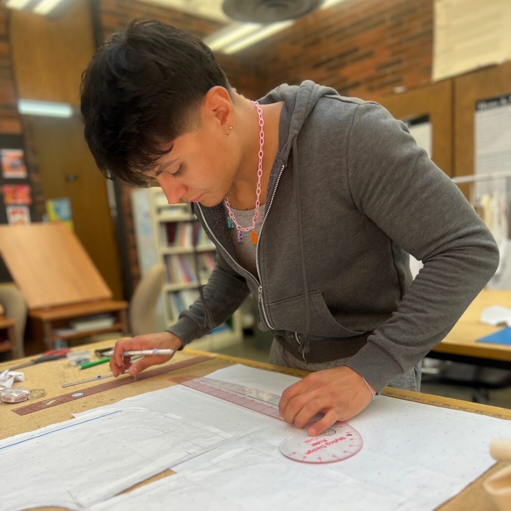 A young fashion designer in a gray hoodie uses a ruler and pencil to draw on pattern paper at a workspace, surrounded by shelves and materials, preparing designs that may one day hit the runway.