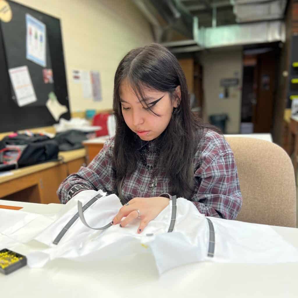A young woman sits at a table concentrating on sewing fabric pieces together in a classroom or workshop setting.