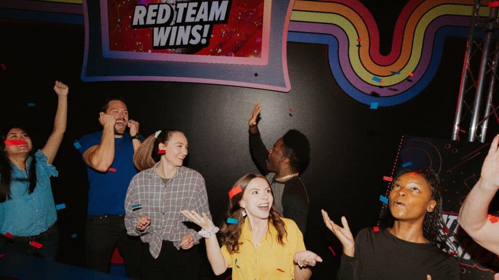 Six people celebrate under a "RED TEAM WINS!" sign, smiling and throwing confetti in a brightly colored room with a retro design.