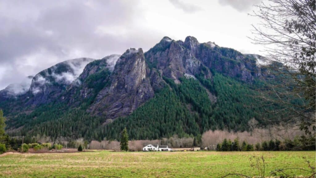 Large rocky mountain with dense green forest at its base, cloudy sky above, open grassy field and a white house in the foreground.