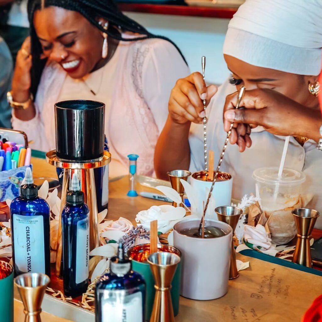 Two women sit at a table making candles, surrounded by bottles, containers, and tools, with one woman stirring wax and the other laughing.