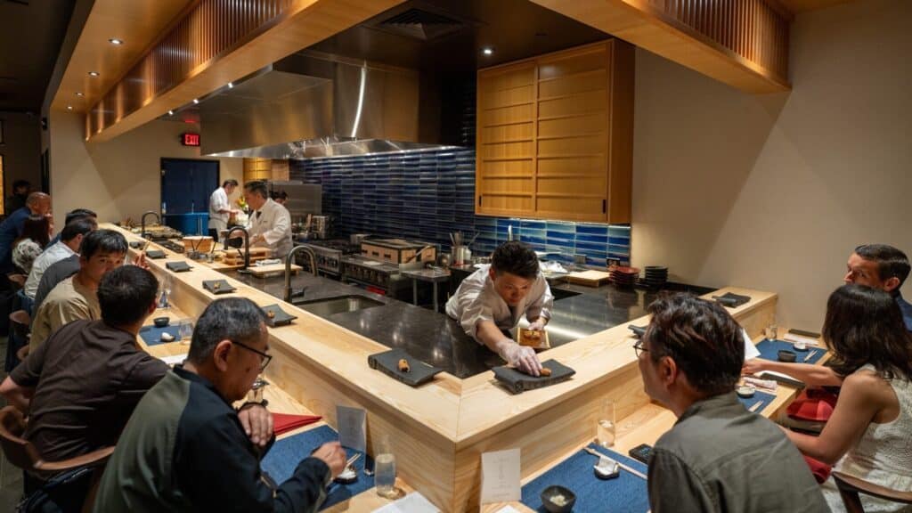 Chefs prepare food behind a wooden sushi bar, serving and interacting with seated diners in a modern, well-lit restaurant.