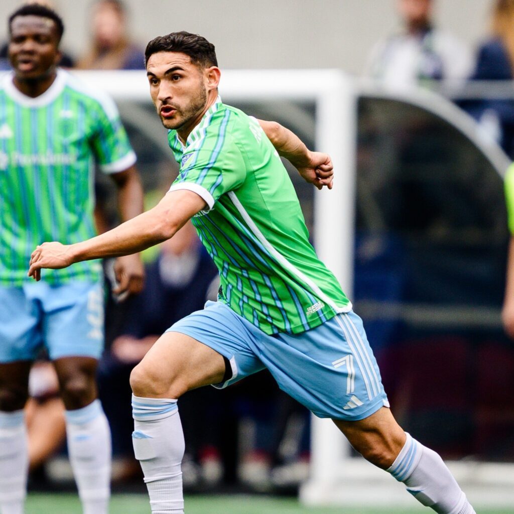 A soccer player in a green and blue striped jersey and blue shorts is running on the field during a match. Another player stands in the background, capturing the energy perfect for a Seattle Mother's Day gift or moment.