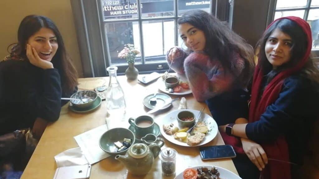 Three women sit at a wooden table in a cafe with plates of food, cups, and a teapot in front of them. Two of them look at the camera; one is smiling, the others have neutral expressions.