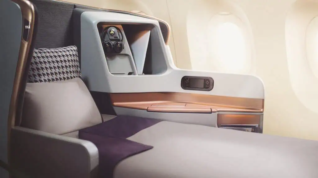 A business class airplane seat fully reclined into a flatbed position, with a pillow and blanket, next to windows and a control panel.