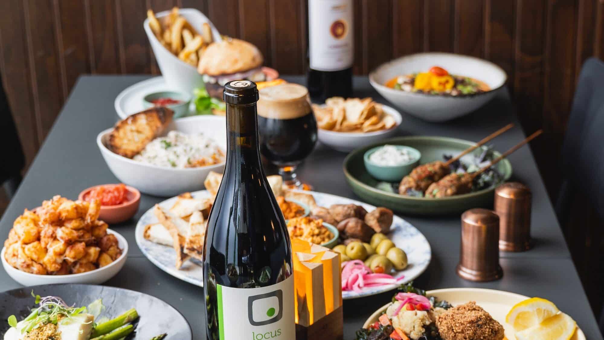 A table is set with various Mediterranean dishes, including olives, pita, grilled skewers, fries, sauces, and bottles of wine and beer.