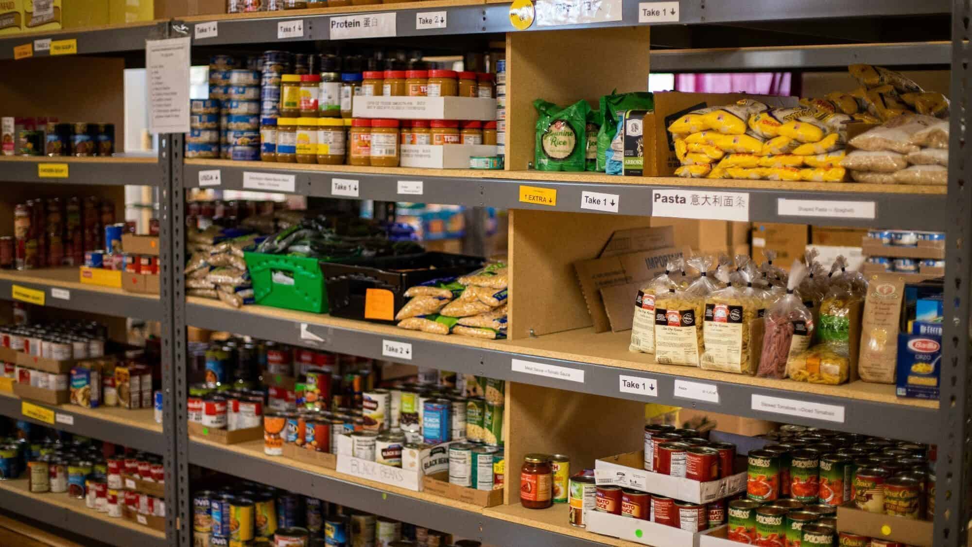 Shelves stocked with canned goods, bags of pasta, rice, beans, and other non-perishable food items in a pantry or food bank.
