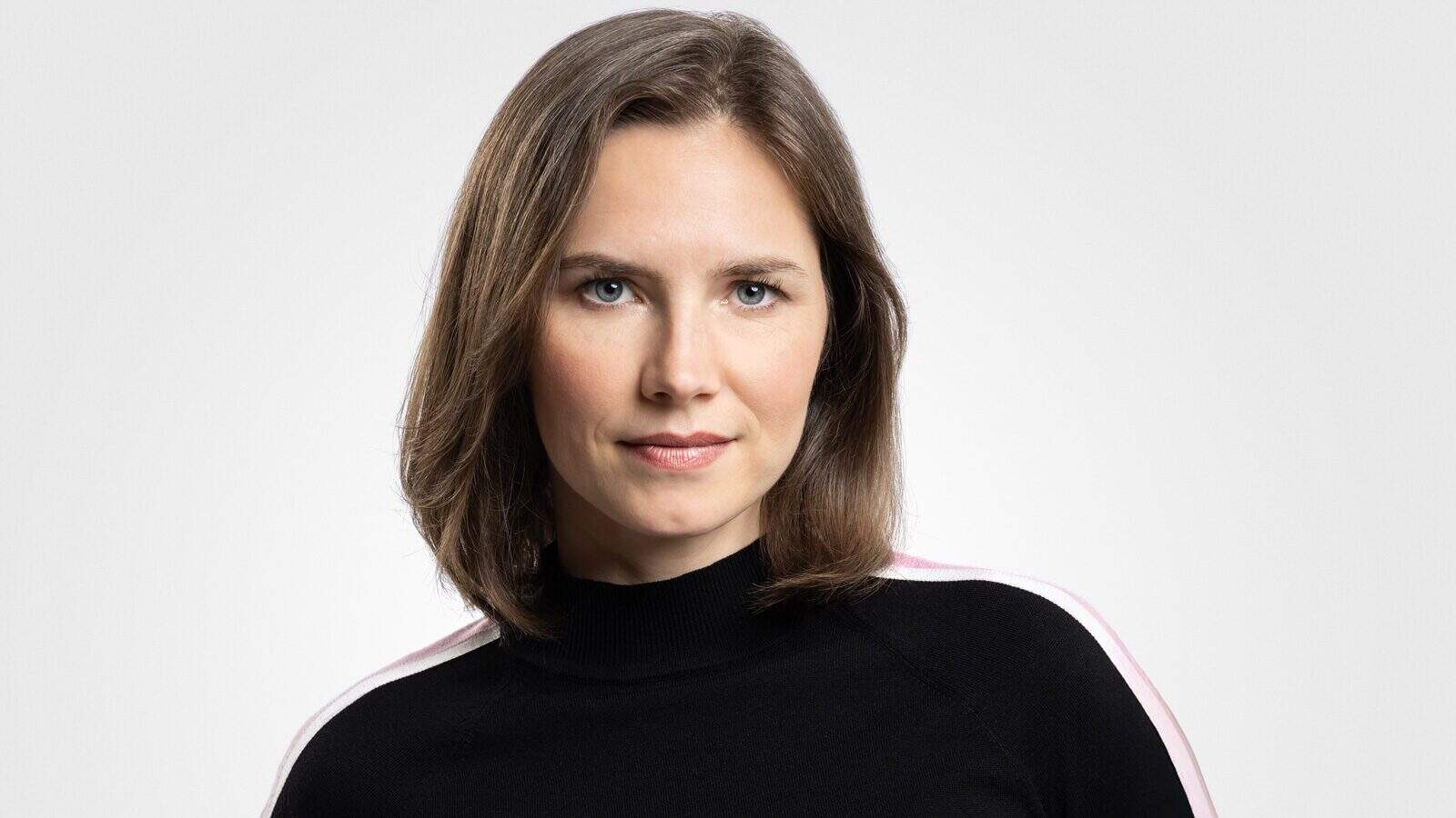 A woman with straight, shoulder-length brown hair and blue eyes wears a black top with white and pink stripes on the shoulders, posing against a plain light background as Amanda Knox works to reclaim her story.
