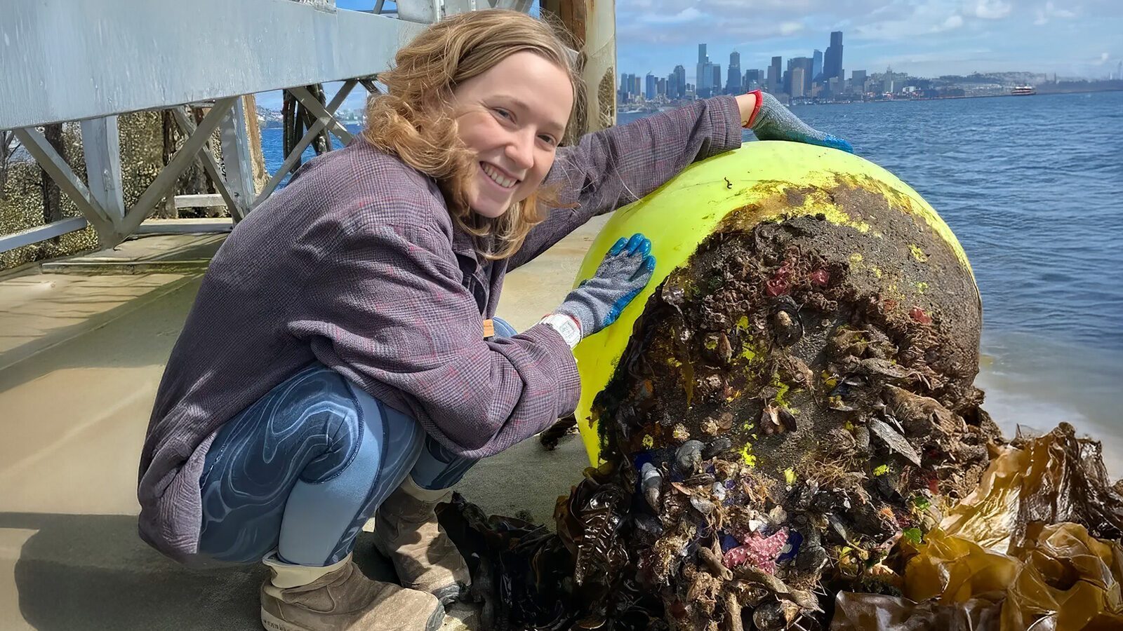 A person crouches beside a barnacle-covered buoy, the city skyline looming in the background. This scene speaks to the important reminder on Earth Day that our connection to nature matters now more than ever.
