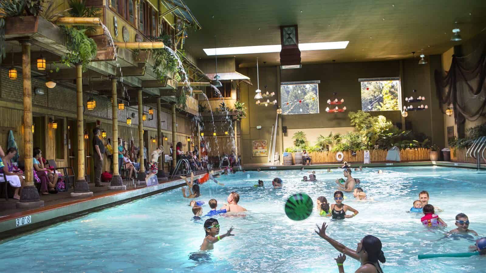 Indoor swimming pool at Anderson School with children and adults playing in the water; poolside seating and tropical-themed decor are visible along the left side—perfect for family fun or swimming lessons.