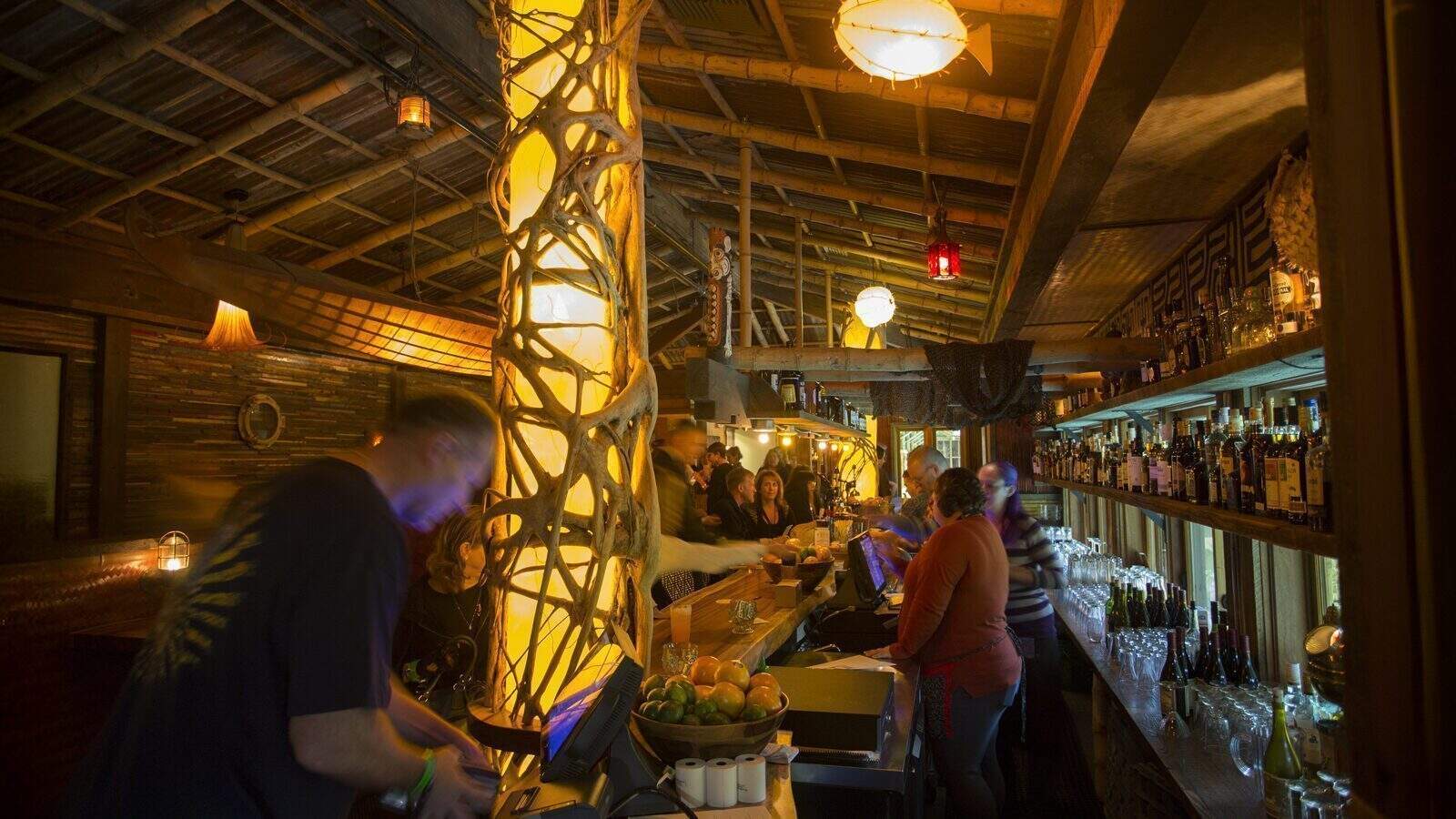 Dimly lit bar at Anderson School with a glowing decorative pillar, bartenders preparing drinks, and patrons seated at the counter, all set within a rustic, wooden interior that celebrates preservation.