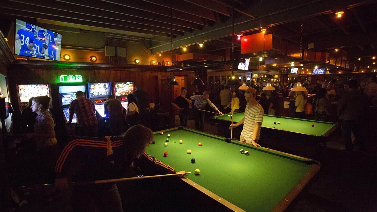 People play pool and arcade games at Anderson School's dimly lit bar, with sports on TVs, neon signs, and multiple pool tables—an inviting spot for fun, friendly competition, and lessons in classic games.