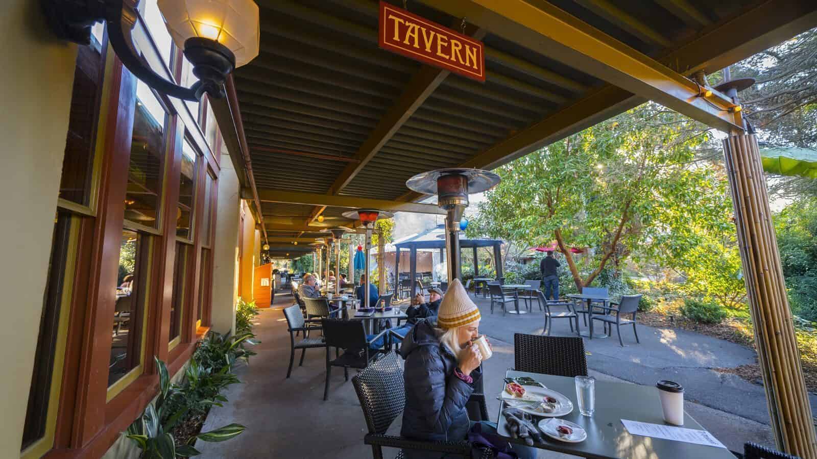 People dine at outdoor tables under a covered patio at Anderson School’s tavern, enjoying food and drinks amid the lush trees—a lively spot where preservation meets relaxation.