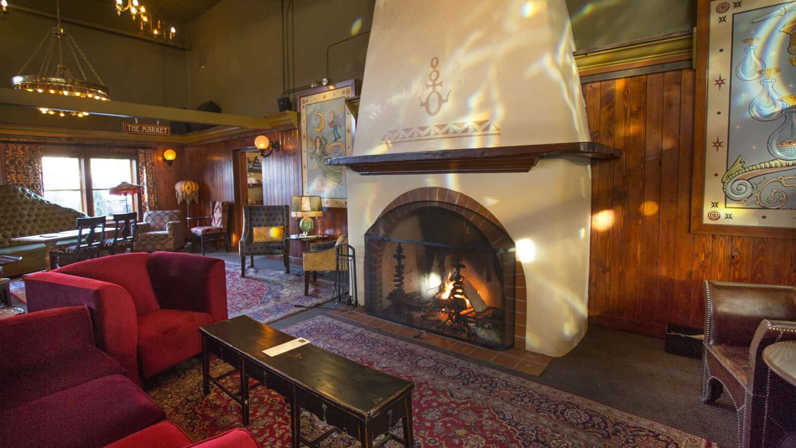 A cozy lounge at Anderson School with red armchairs, wooden tables, and a lit fireplace, decorated with wall art and a patterned rug—a warm space where preservation and comfort blend seamlessly.