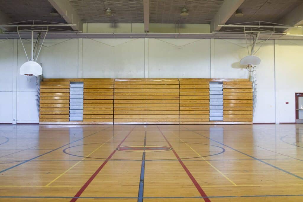 The empty indoor gymnasium at Anderson School features closed wooden bleachers, two basketball hoops, and marked court lines on a polished floor—a space reflecting careful preservation and ready for future lessons.