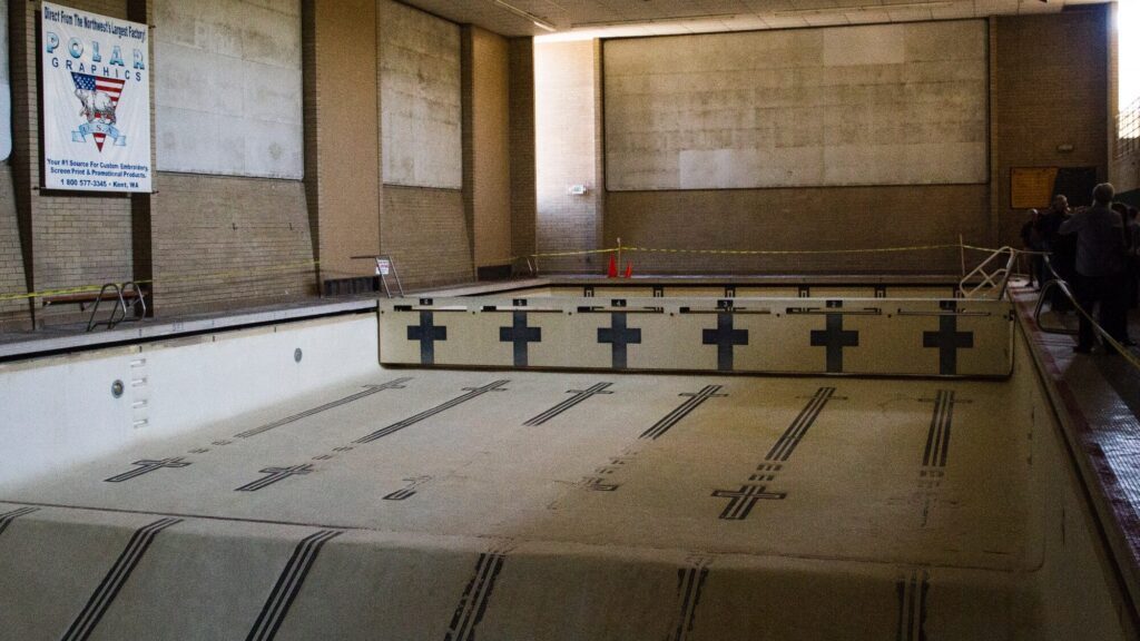 An empty indoor swimming pool with lane markings, bordered by caution tape, sits in a dimly lit, tiled room at Anderson School—preservation underway before new lessons can begin.