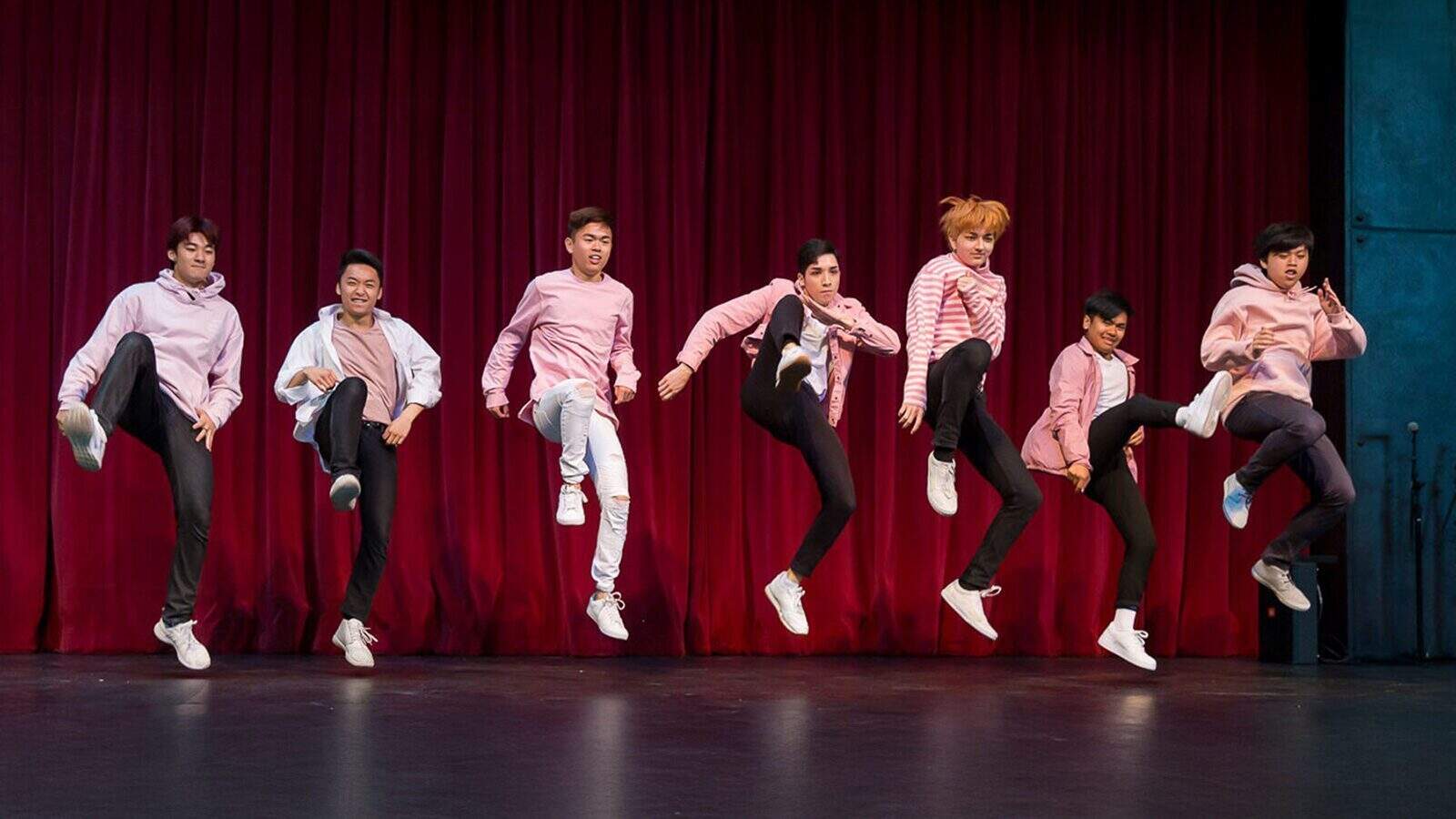 Seven people wearing pink and white outfits perform a synchronized jump with one leg raised in front of a red stage curtain at Sound To Summit: AANHPI Voices Rising in Seattle.