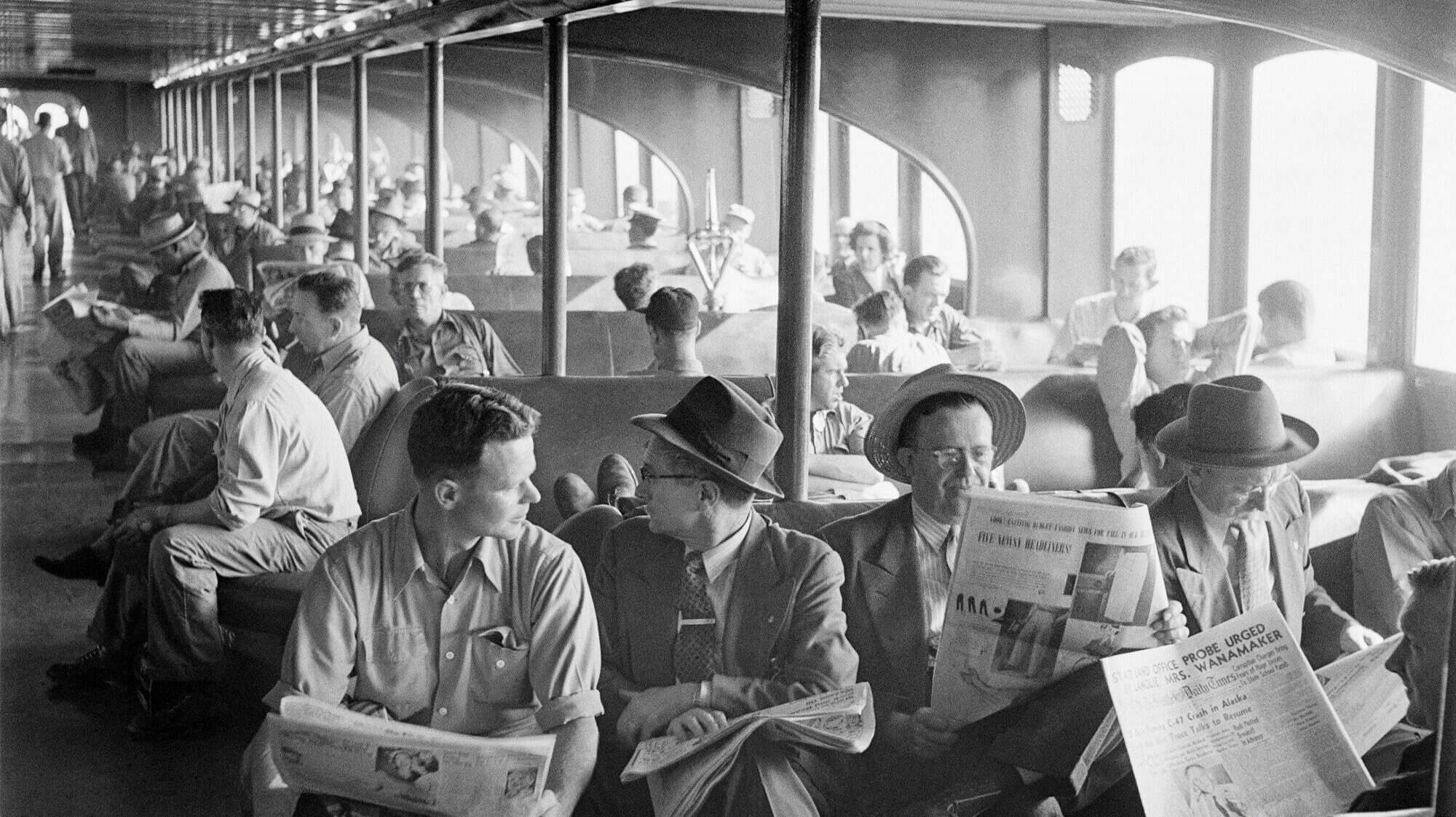 Passengers seated on the Kalakala ferry read newspapers and converse. The space is crowded, with men mostly in hats, shirts, and ties. Artifacts of a bygone era surround them as sunlight streams through the windows on one side.