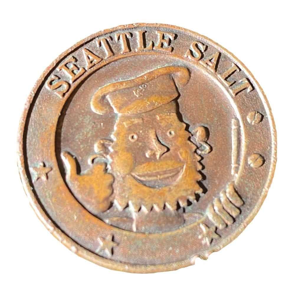 A round, weathered metal coin featuring a bearded sailor giving a thumbs-up, reminiscent of the lively spirit aboard the historic Kalakala ferry. Text reads "SEATTLE SALT.