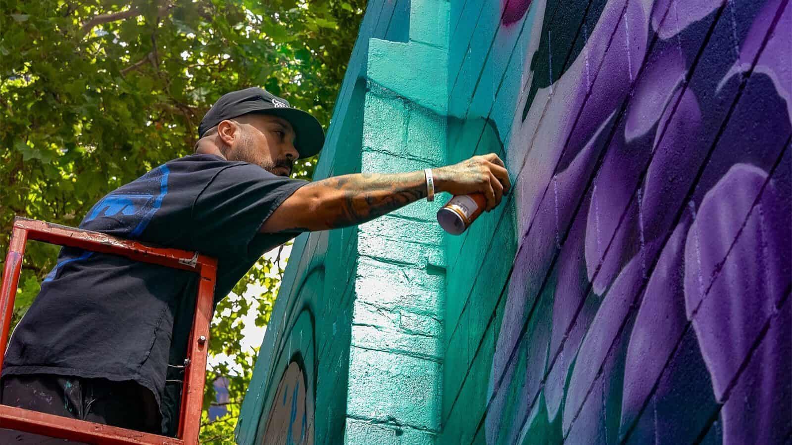 A person on a ladder spray paints a colorful mural on a brick wall outdoors in Seattle, celebrating Hispanic Heritage Month, with trees visible in the background.