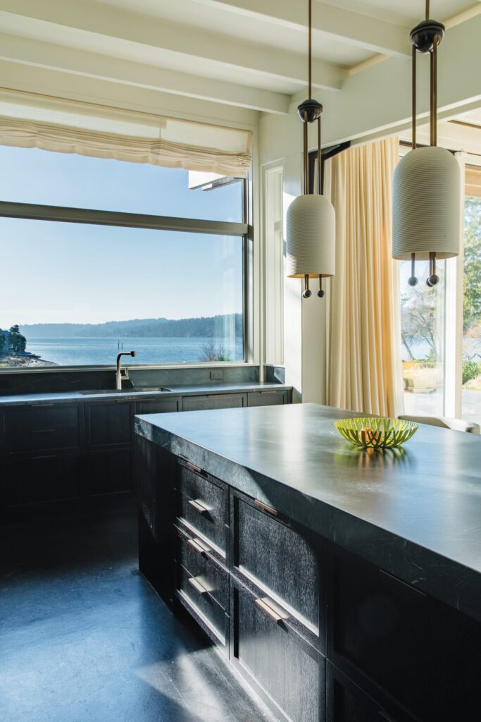 A modern kitchen with dark cabinetry and a large island, perfect for those dreaming of retiring on the beach. Pendant lighting illuminates a fruit bowl, while beige curtains frame a large window with serene lake views—a delightful second home retreat.