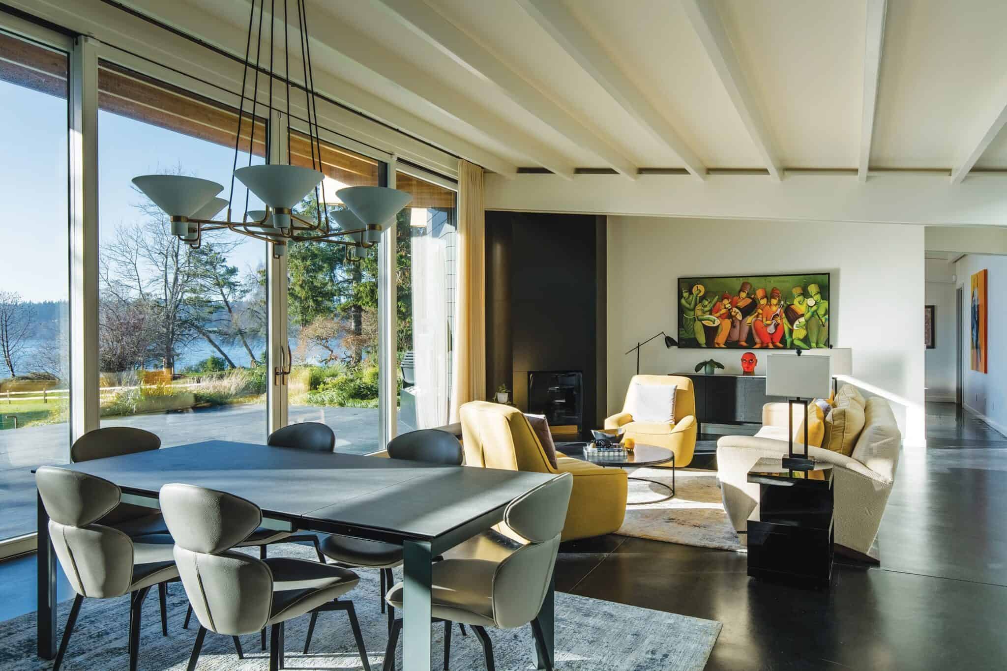 A modern living and dining area in what could be the ideal second home, featuring large windows, a beige sofa, yellow chairs, and a dark dining table with gray chairs. A colorful painting adorns the wall while offering a scenic view of trees and water outside.