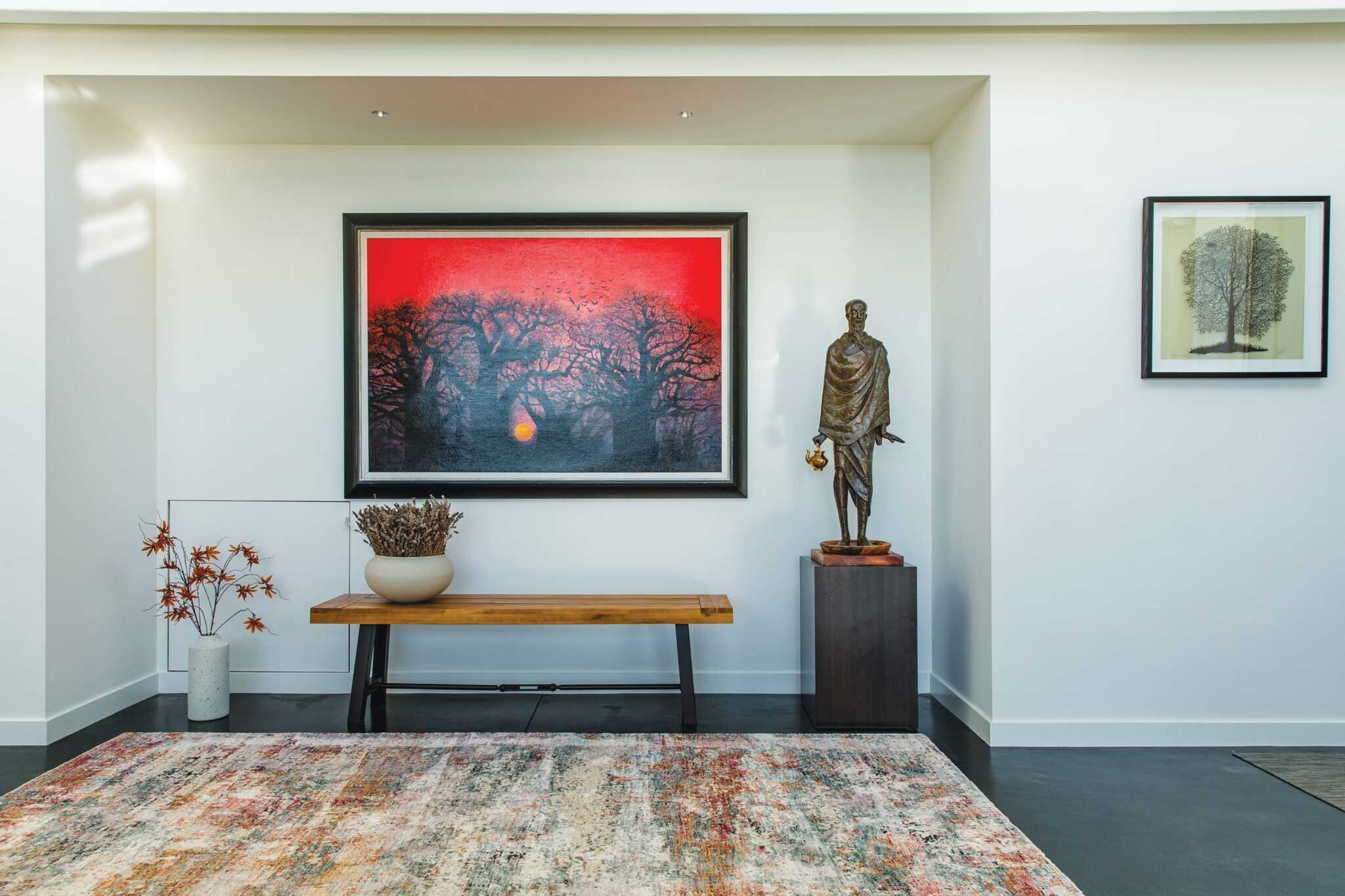 The room, perfect for second homes, features a red tree painting, a statue, two framed artworks, a bench on an inviting rug, lush potted plants, and pristine white walls.
