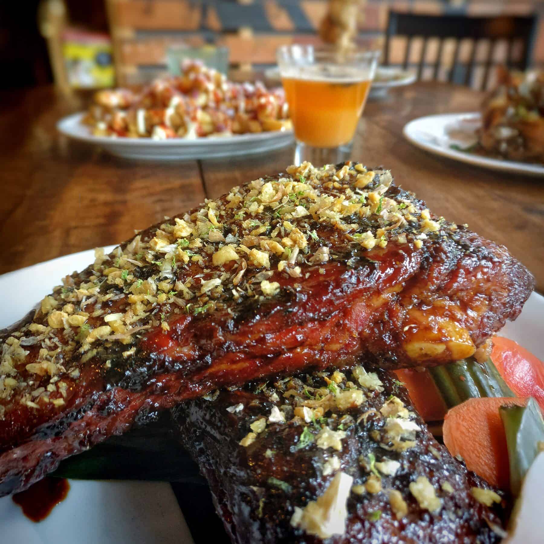 Close-up of glazed ribs topped with herbs and chopped garlic, served with vegetables on a plate; perfect for fall recipes and seasonal eating. A glass of beer and other dishes are in the background.