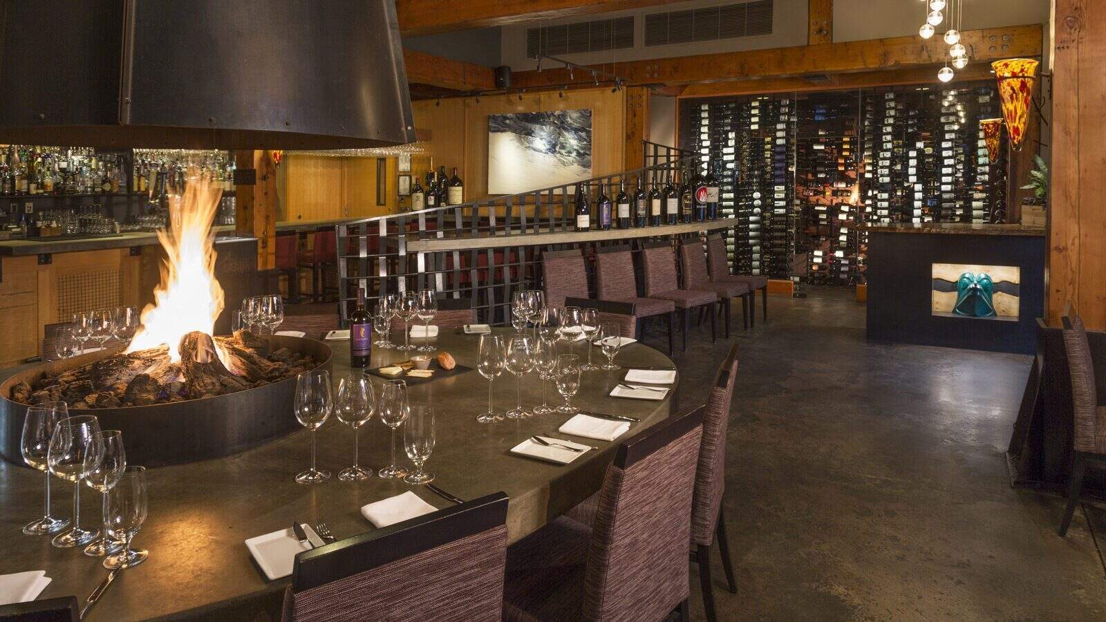 A modern Woodinville restaurant dining area with a round table set for guests, a central fireplace, a stunning wine display wall, and contemporary decor designed for ultimate relaxation.