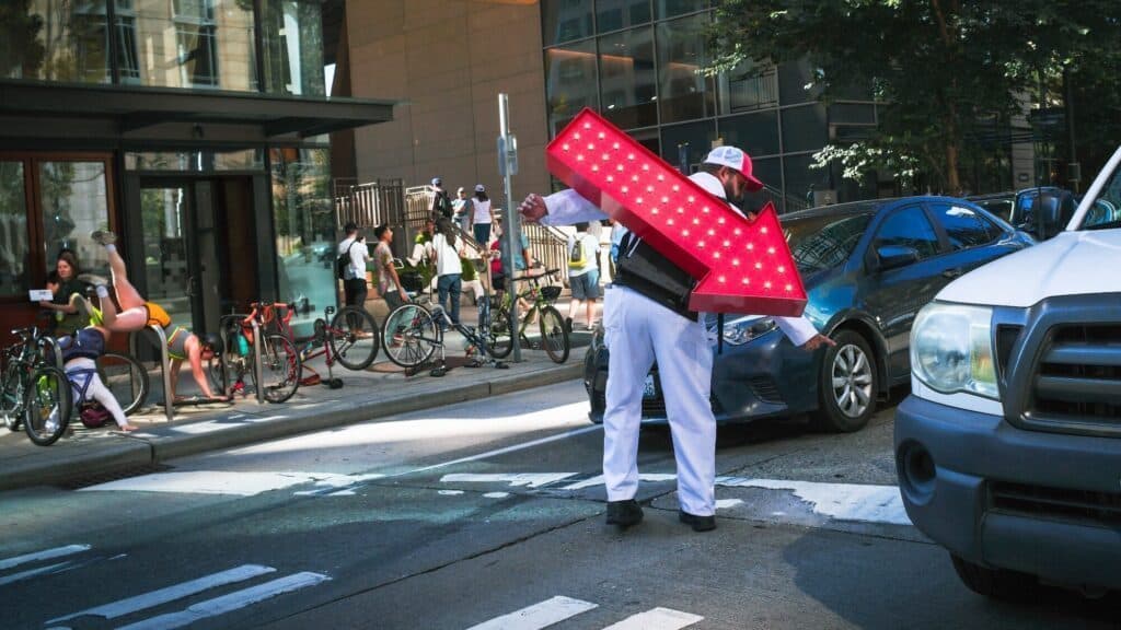 A person in white clothing and a cap directs traffic with a large, illuminated red arrow sign on a city street, as cars and cyclists move nearby.
