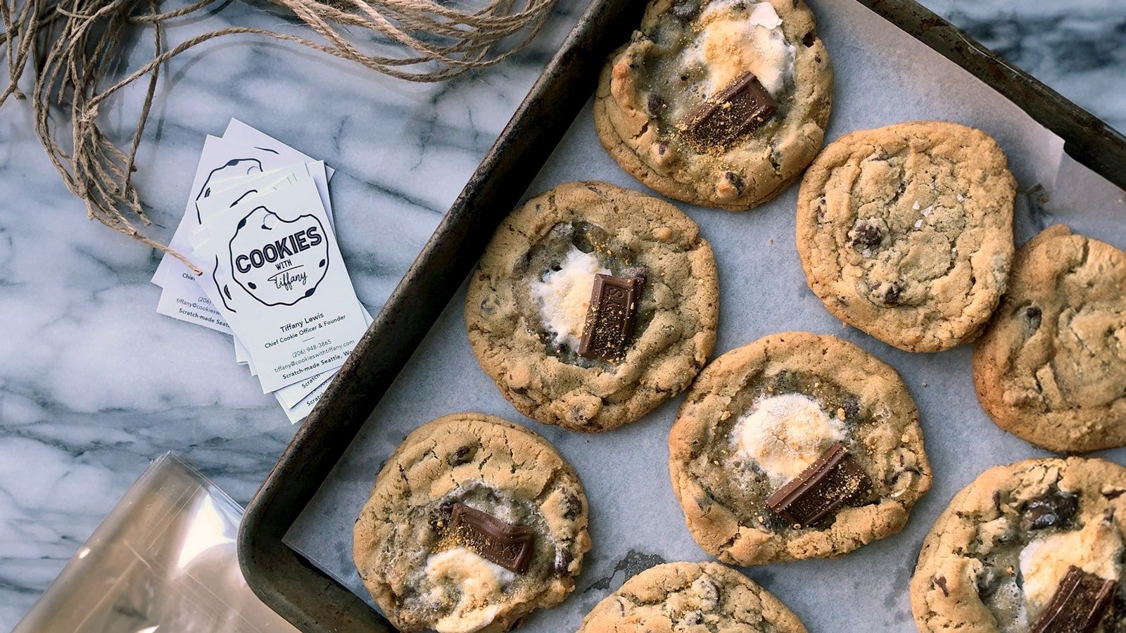 A baking sheet with eight chocolate chip cookies topped with marshmallow and chocolate pieces rests on a marble surface, reminiscent of Seattle restaurants' innovative dessert offerings, accompanied by some string and cookie-themed cards.