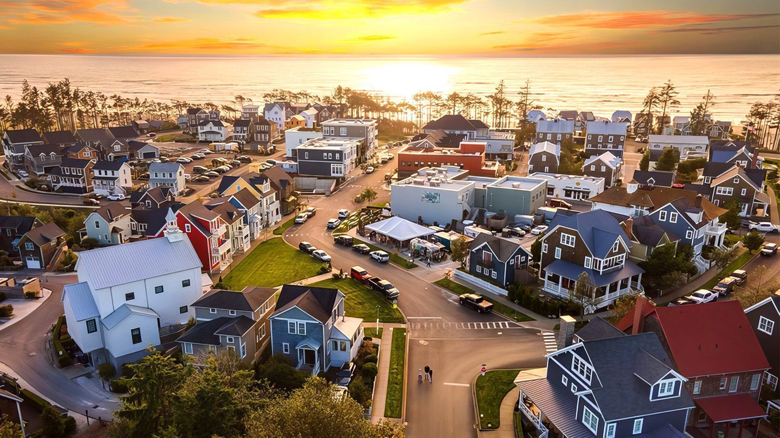 A coastal residential neighborhood at sunset, featuring a variety of colorful houses, trees, and parked cars along winding streets with the picturesque Pacific coastline in the background.