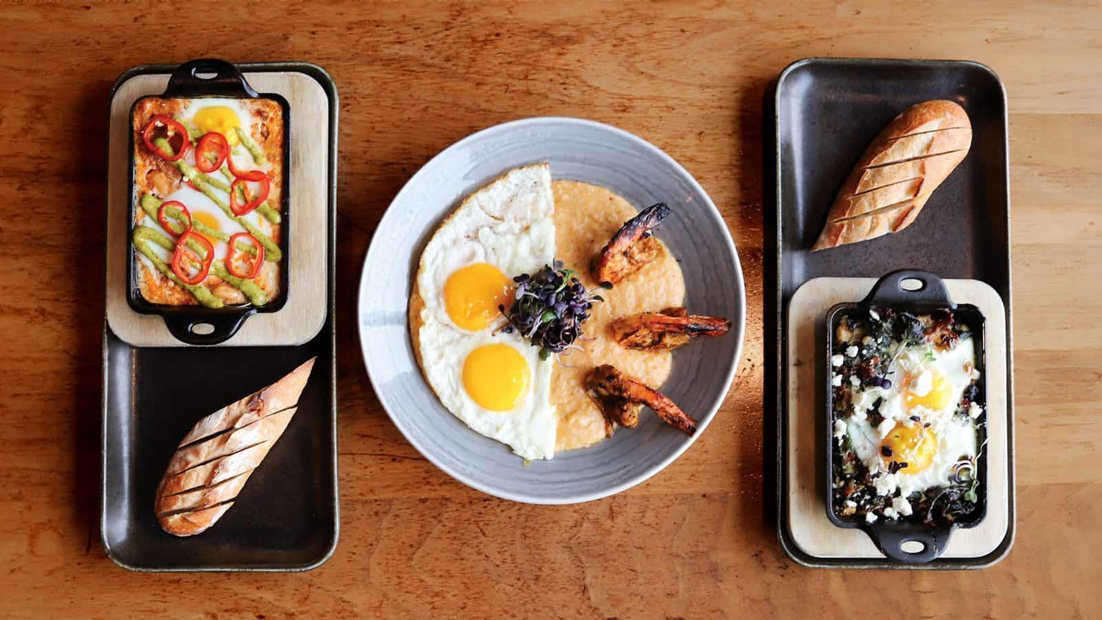A delightful brunch experience awaits with dishes of baked eggs topped to perfection, eggs over shrimp and grits, and sliced bread on trays, all set beautifully on a wooden table. Discover this culinary gem among Seattle restaurants.