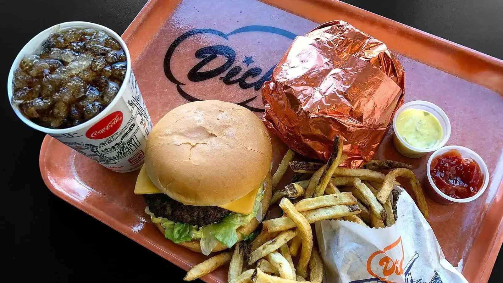 A tray featuring a wrapped burger, an unwrapped cheeseburger, fries, soda, and small cups of mustard and ketchup awaits your discovery at one of the exciting new Seattle restaurants.