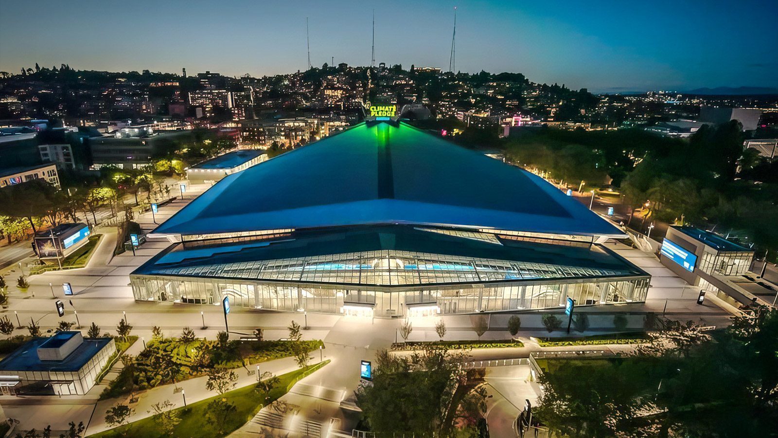 Aerial view of a large, illuminated triangular-roofed concert venue at night, surrounded by trees and city lights in the background.