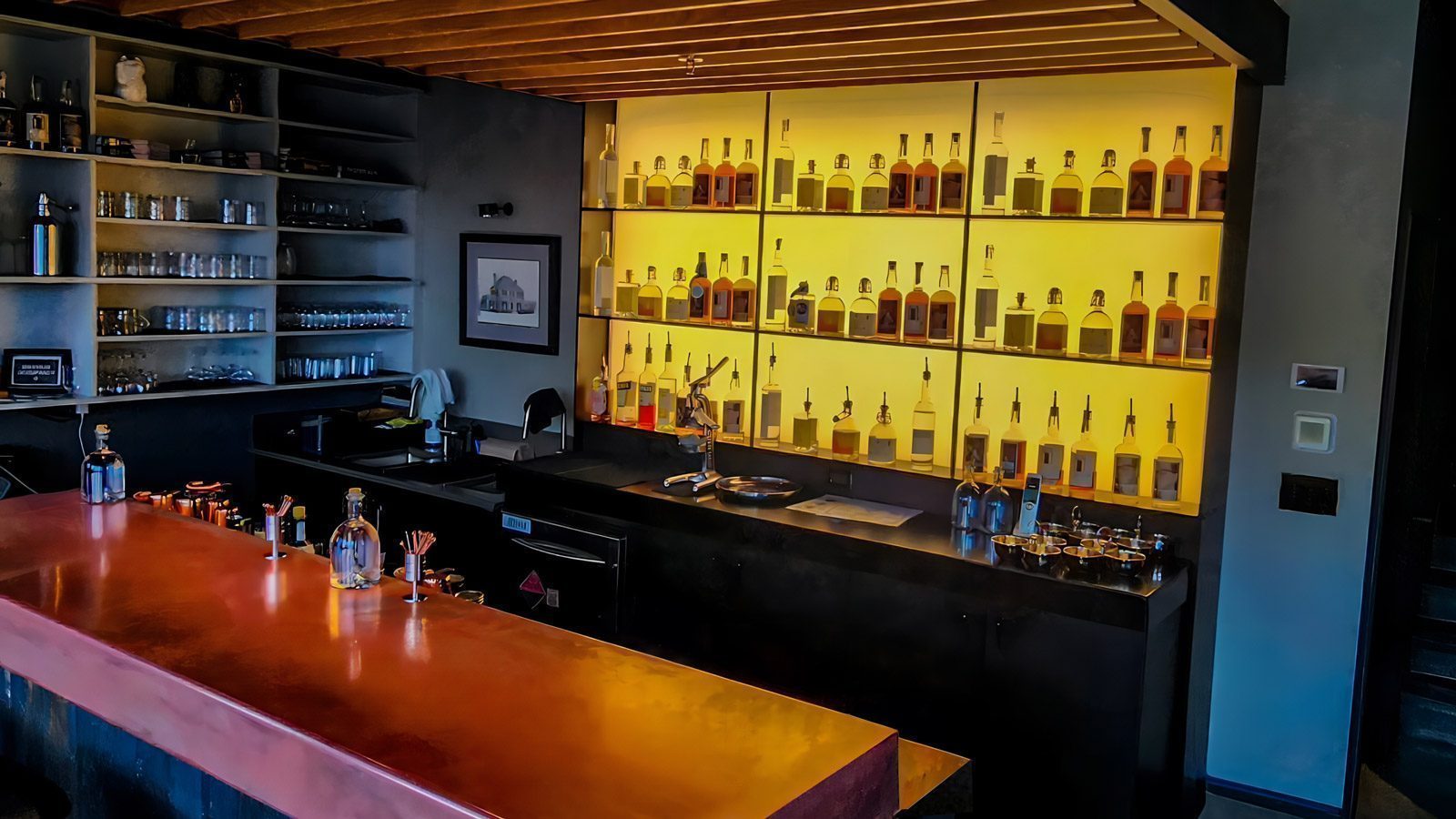 Modern bar with a red countertop, shelves of glasses, and illuminated shelves displaying bottles from Wildwood Spirits Co., resembling a chic distillery setting.