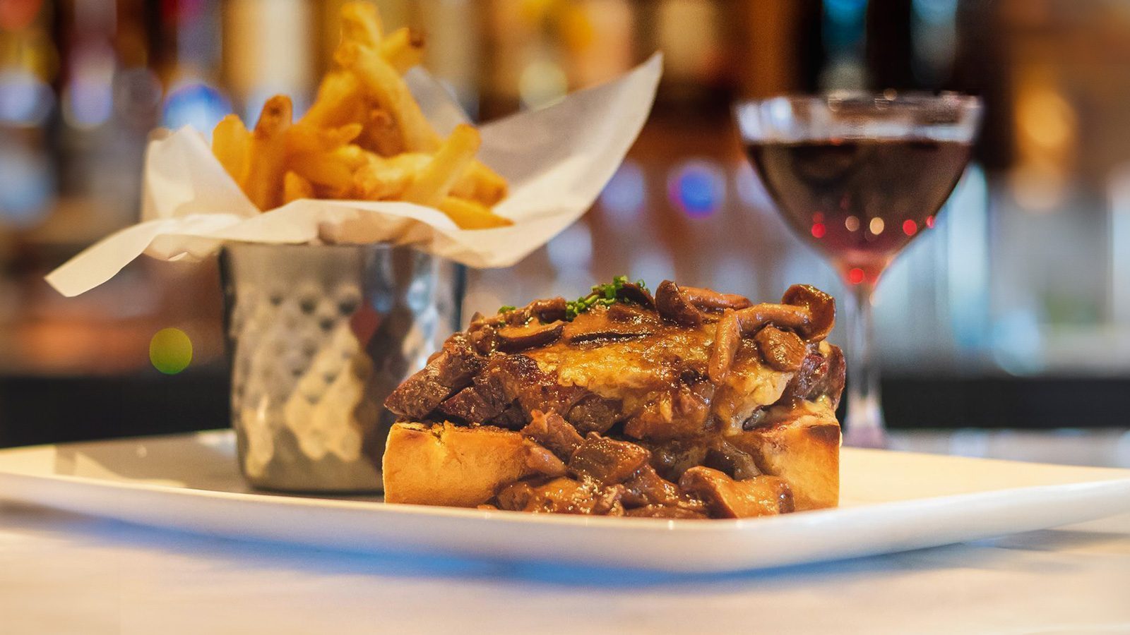 Enjoy an open-faced sandwich with beef and mushrooms on toasted bread, paired perfectly with fries in a metal cup and a glass of red wine at John Howie Steak's happy hour, all elegantly presented on a white plate.