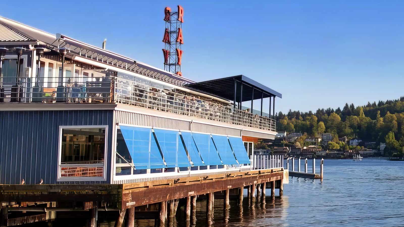 A restaurant with large blue windows on a pier extends over the water, offering outdoor dining with Olympic Mountains views. A neon sign reads "RAY'S." Trees and houses are visible in the background under a clear blue sky.