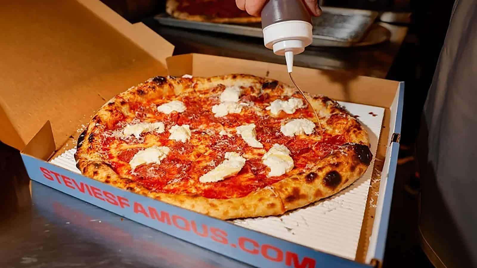 A pizza in a delivery box is topped with dollops of white cheese, while a sauce is expertly drizzled over it from a bottle—a signature touch from one of the exciting new Seattle restaurants.