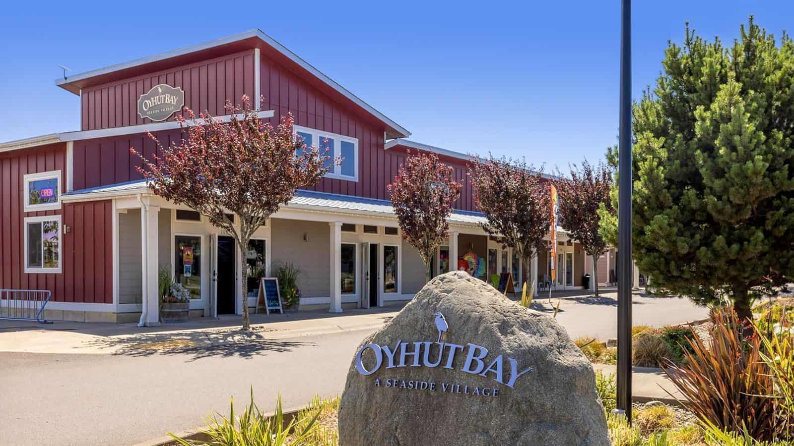 The Oyhut Bay building, adorned with red siding, sits near a large stone sign, welcoming visitors. Trees line the walkway under a clear blue sky, offering easy access to the nearby Oyhut Bay Wildlife Reserve and various lodging options.