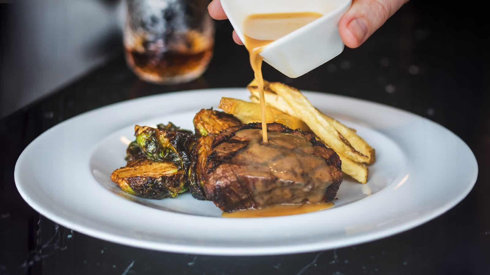 A plate at John Howie Steak features a delectable combination of steak, Brussels sprouts, and fries. A rich sauce is being poured over the tender steak, enhancing each bite. This presentation showcases why it’s known as one of the best restaurants around.