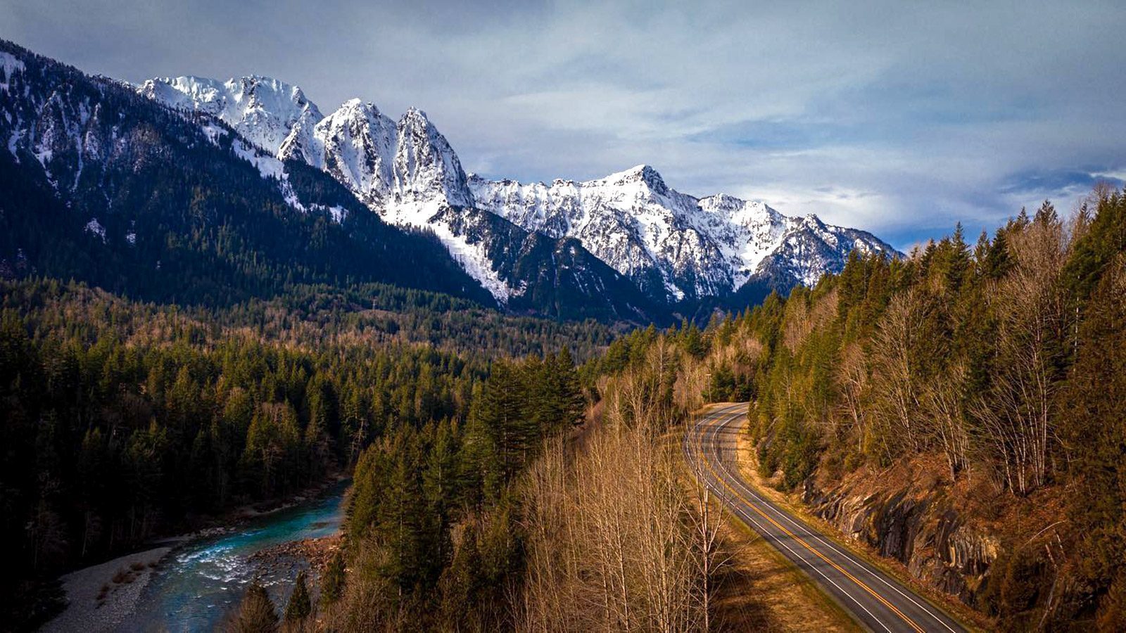 A winding road invites travelers on a Cascade Loop adventure through a forest, with snow-capped mountains standing majestically in the background under a cloudy sky.