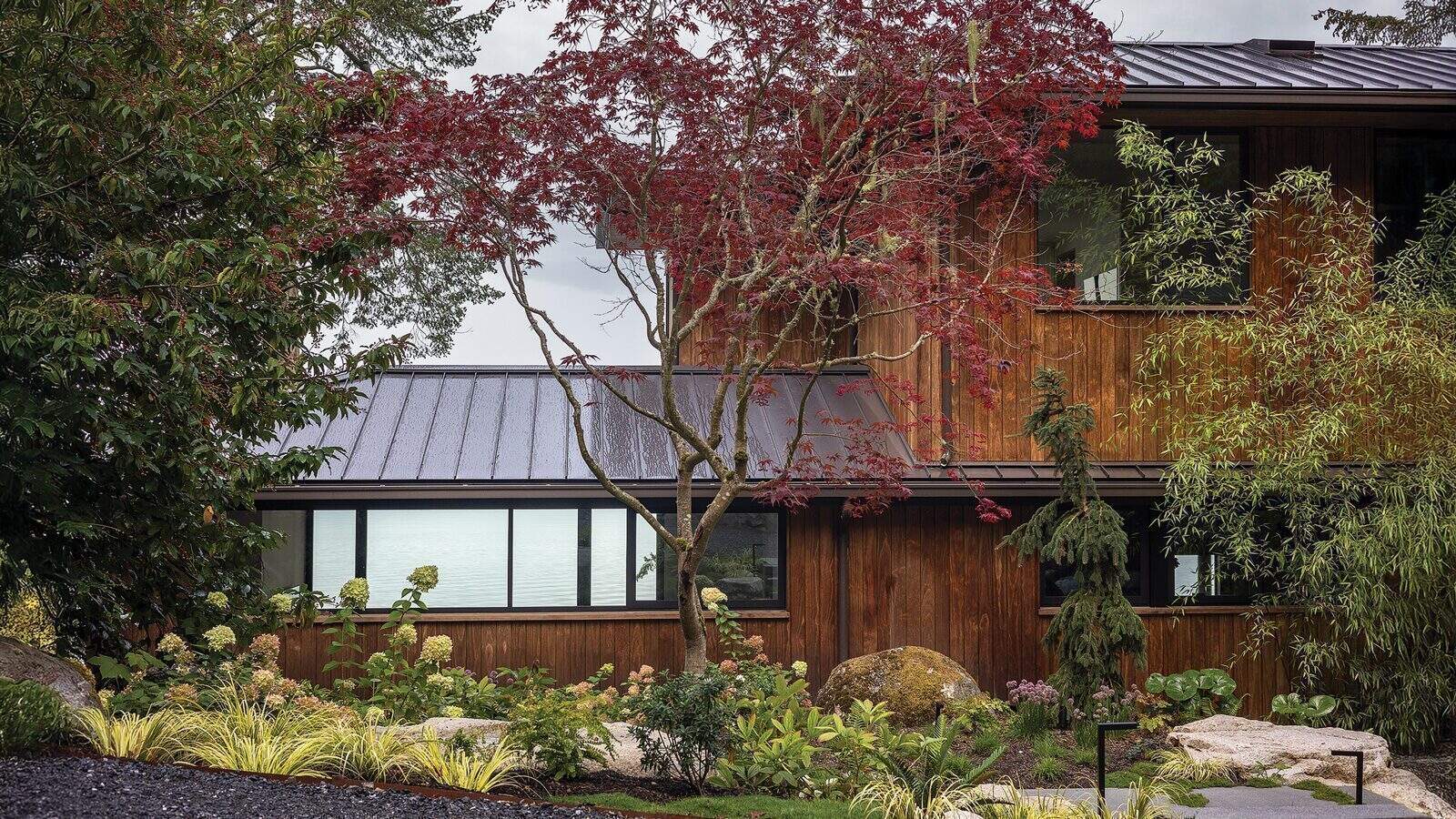 Two-story house with wooden exterior, large windows, and metal roof on Orcas Island, surrounded by lush garden plants, trees, and large rocks—perfect for a serene home renovation.