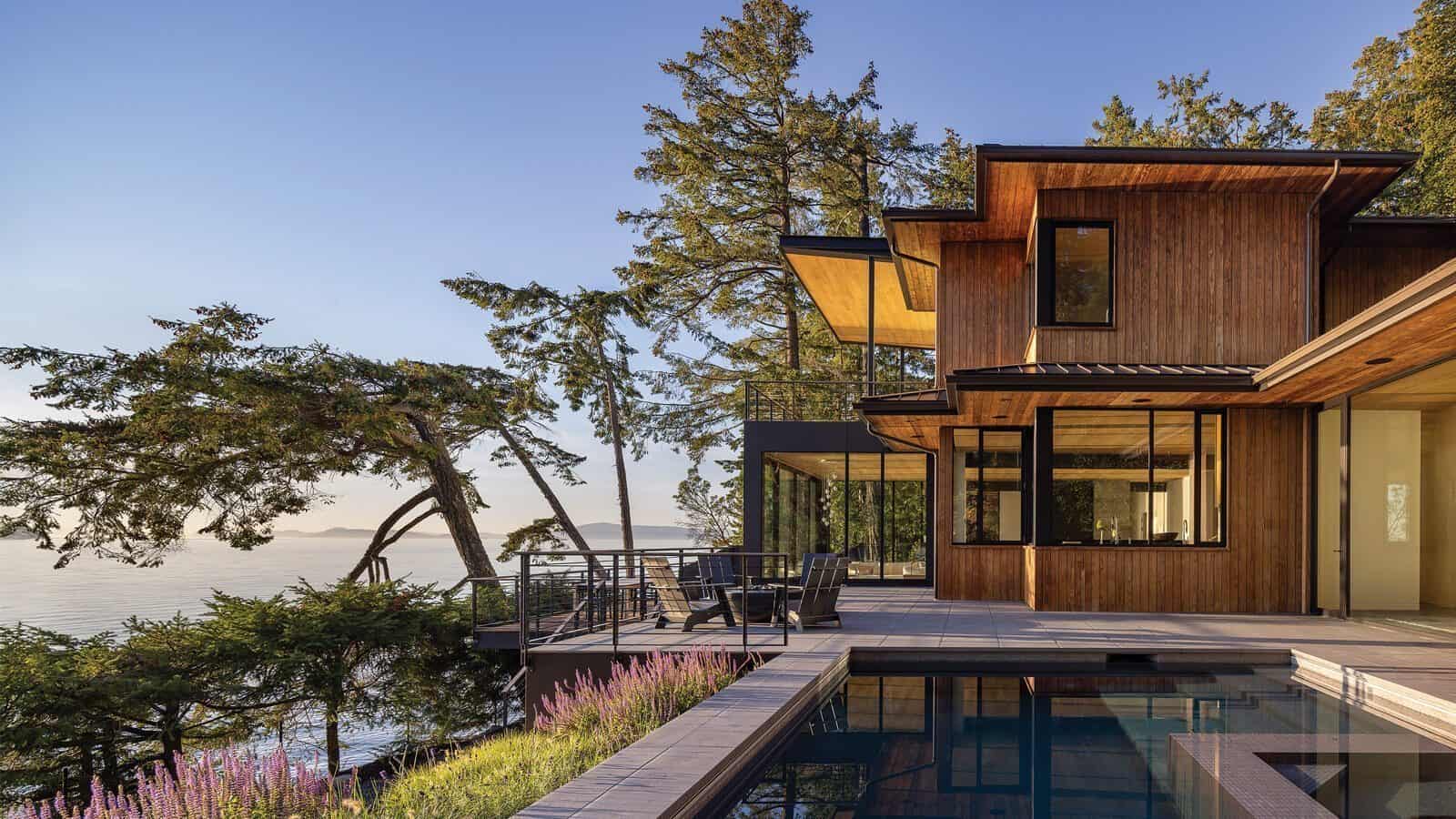 A modern wooden house on Orcas Island with large windows overlooks a pool and outdoor seating area, surrounded by trees and facing a waterfront view.