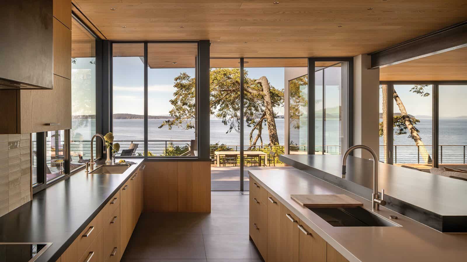 Modern kitchen with light wood cabinets and large windows offering a view of trees, patio furniture, and a body of water outside—a serene Orcas Island retreat perfect for your next home renovation inspiration.