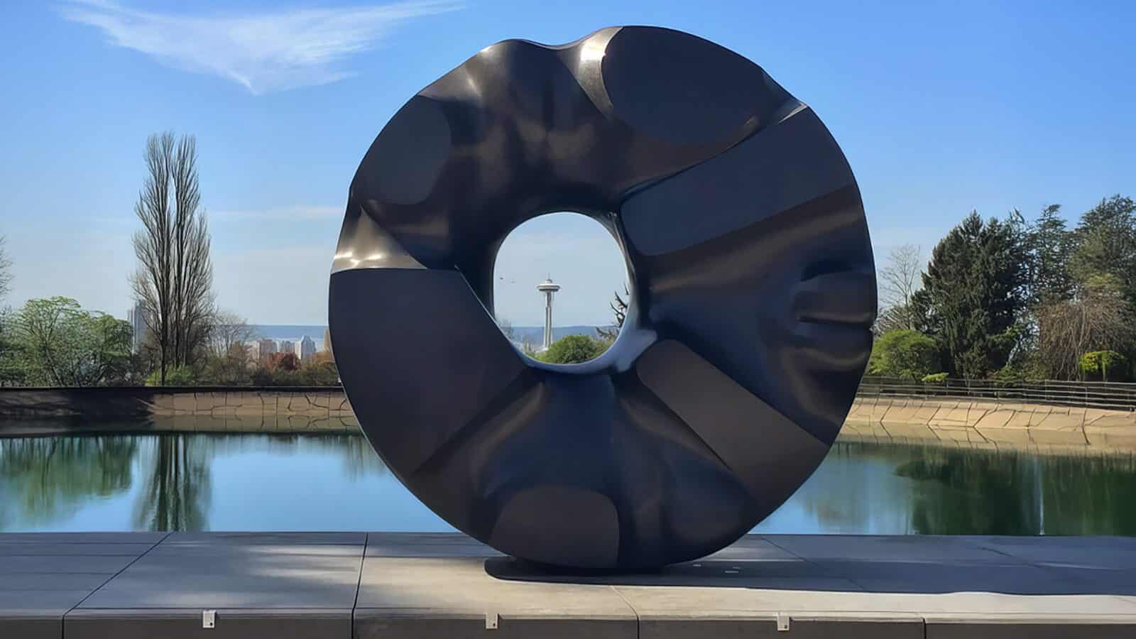 Large black abstract circular sculpture with a central hole, displayed outdoors near a reflective pool, stands among trees and a tower—one of the striking Seattle cultural landmarks.